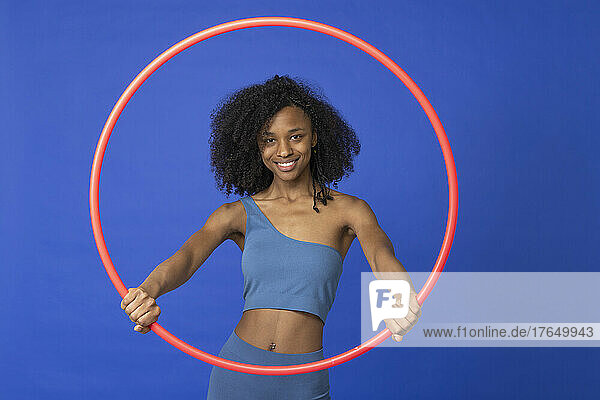 Smiling young woman holding red hoop standing against blue background
