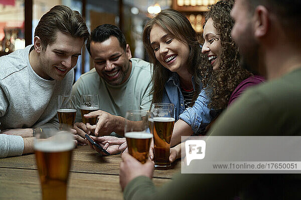 Gruppe glücklicher Freunde  die in einer Kneipe Bier trinken und ein Smartphone teilen