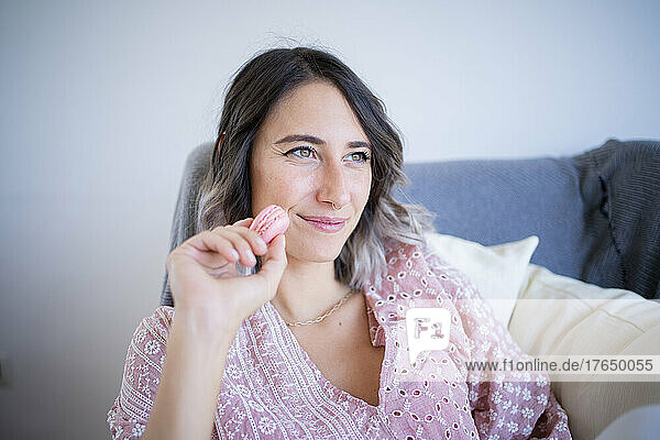 Thoughtful smiling woman with macaroon sitting in sofa at home