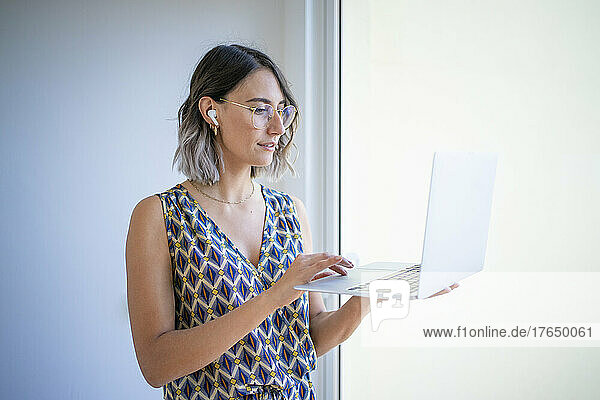 Young freelancer working on laptop by glass window at home
