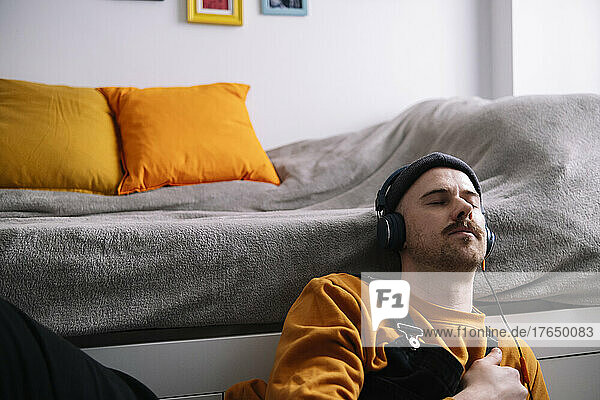 Man with eyes closed listening music through headphones sitting by bed at home