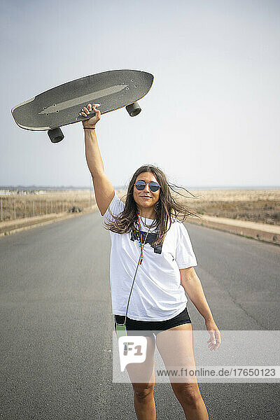 Smiling woman with sunglasses holding skateboard aloft on road