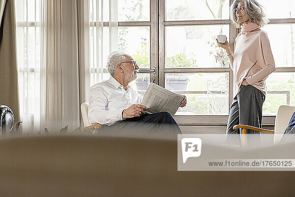 Smiling woman holding coffee cup talking with man holding newspaper in living room at home