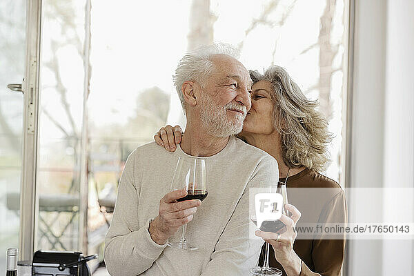 Ältere Frau hält Weinglas und küsst Mann vor Fenster in Hotelwohnung
