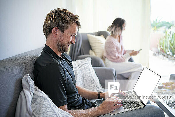 Happy freelancer working on laptop with woman using smart phone in living room