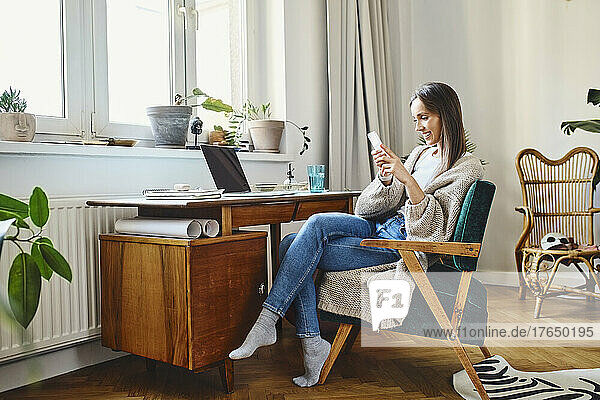 Smiling freelancer using mobile phone sitting on chair at home