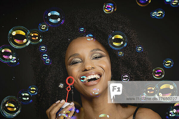 Happy young woman holding bubble wand amidst bubbles against black background