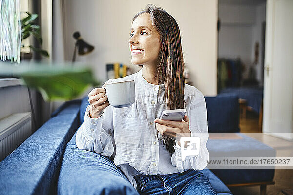 Smiling woman with coffee cup and smart phone sitting on sofa at home