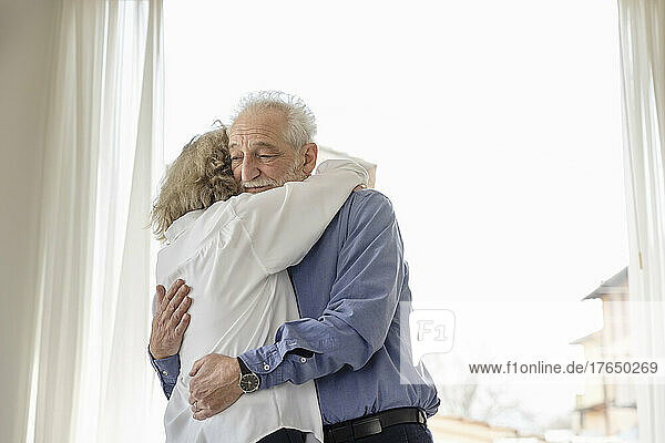 Senior couple hugging each other standing by window at home