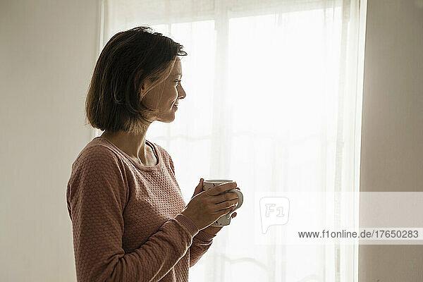 Lächelnde Frau mit Kaffeetasse  die zu Hause am Fenster steht
