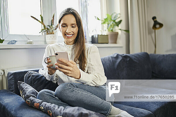 Happy woman surfing net through smart phone sitting on sofa at home