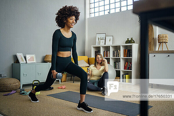 Smiling young woman with dumbbells doing lunges by friend sitting in living room