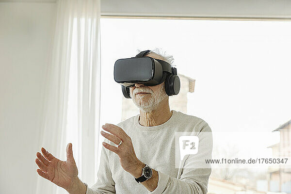 Senior man wearing virtual reality simulator gesturing standing in front of window