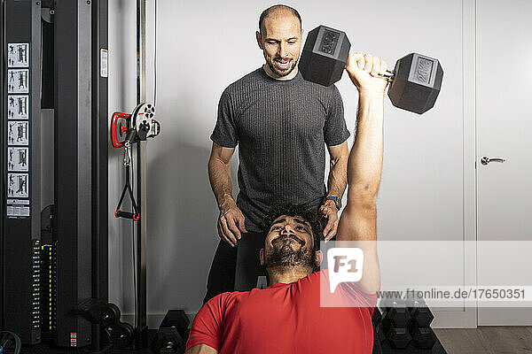 Fitness instructor motivating young man lifting dumbbell at gym