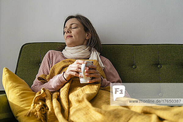 Sick woman holding tea cup resting on sofa at home