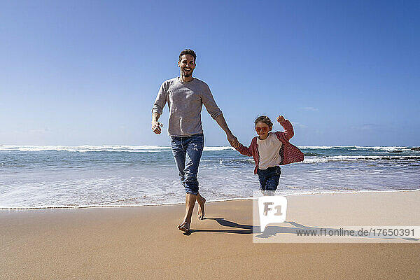 Happy father and daughter holding hands running at beach on sunny day