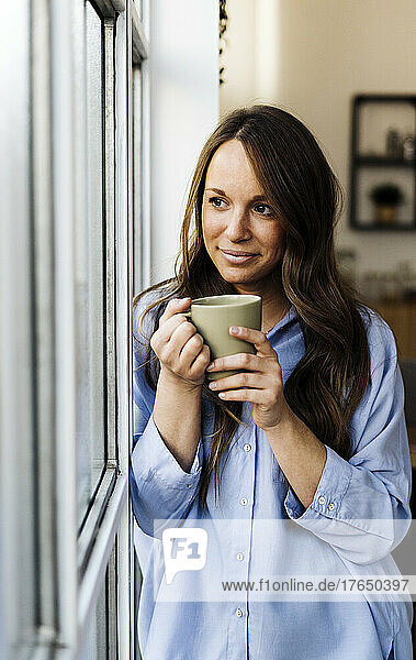 Smiling beautiful woman with coffee cup standing by window at home