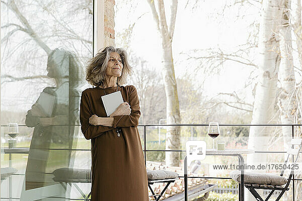 Smiling senior woman with book leaning on glass window in balcony
