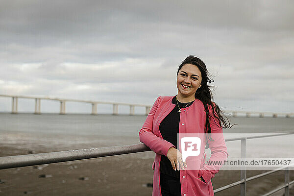 Lächelnde Frau mit der Hand in der Tasche steht am Geländer vor dem Meer