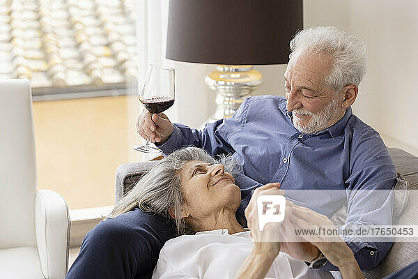 Smiling Senior woman lying on husband's lap sitting with wineglass on sofa at home