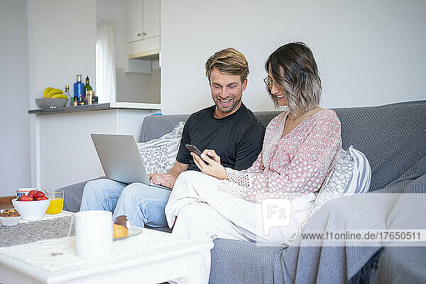 Happy freelancer with laptop looking at woman using smart phone on sofa at home