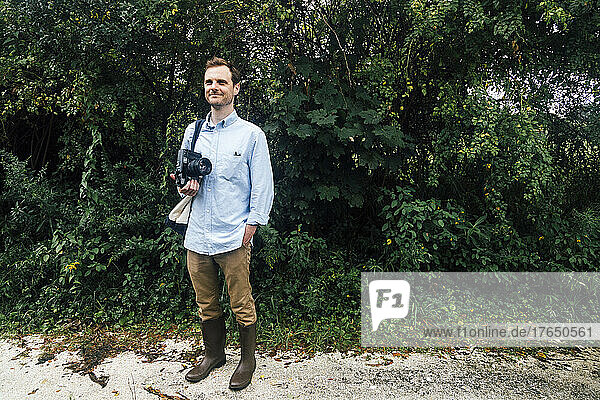 Smiling man holding vintage medium format camera standing with hand in pocket at park