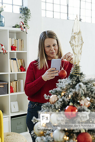 Smiling woman photographing bauble with mobile phone in front of Christmas tree at home