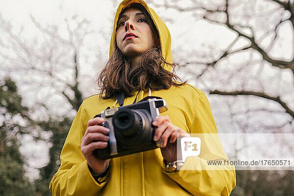 Young woman wearing yellow raincoat standing with camera in forest
