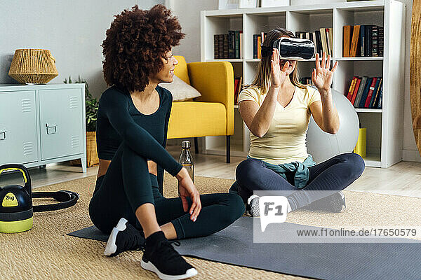 Young woman looking at friend wearing virtual reality simulator in living room at home