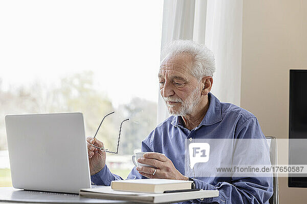 Senior man with laptop and coffee cup sitting by window at hotel apartment