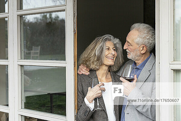 Smiling senior couple with wineglasses standing at doorway