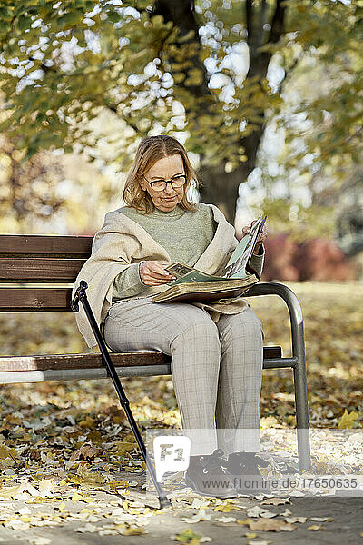 Ältere Frau schaut sich Fotoalbum an und sitzt mit Gehstock auf Bank im Park