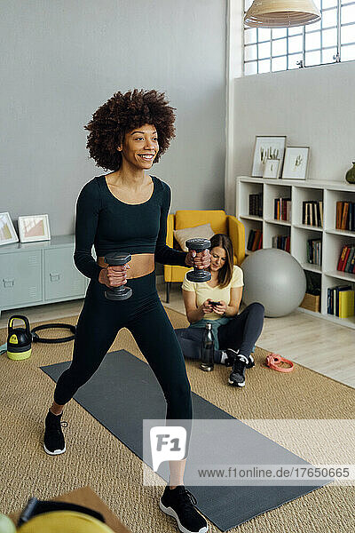 Smiling woman exercising with dumbbell by friend using smart phone in living room