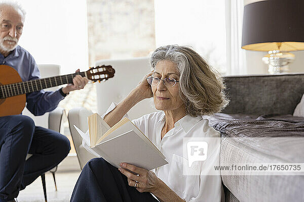 Senior woman with hand in hair reading book by man playing guitar at home