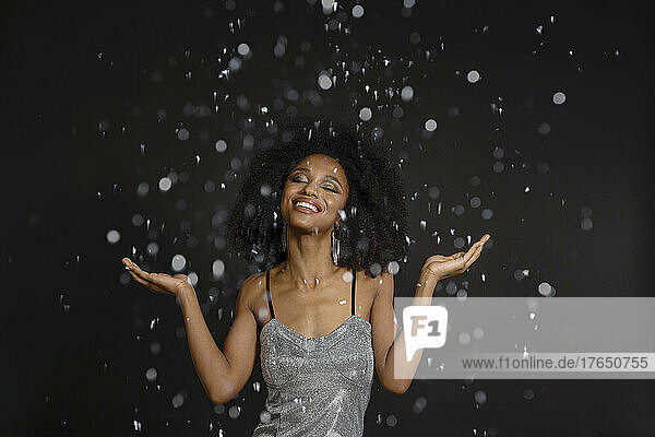 Smiling young woman with eyes closed standing under falling confetti against black background