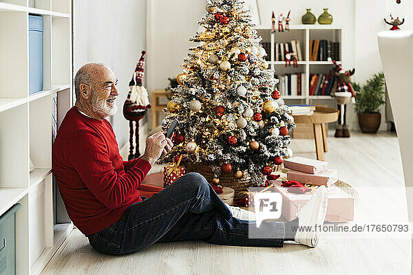 Fröhlicher älterer Mann mit Smartphone sitzt zu Hause am Weihnachtsbaum