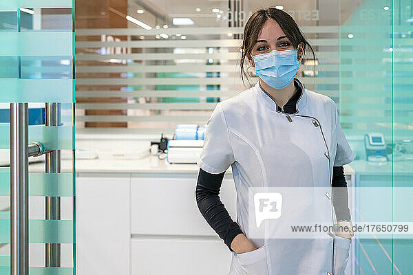 Nurse in protective facial mask standing with hands in pockets at hospital