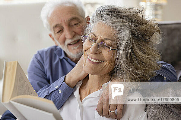 Älterer Mann umarmt Frau  die zu Hause im Wohnzimmer ein Buch liest