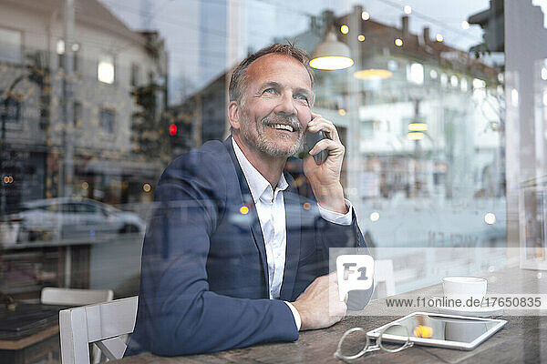 Smiling businessman talking on mobile phone and looking through window at cafe