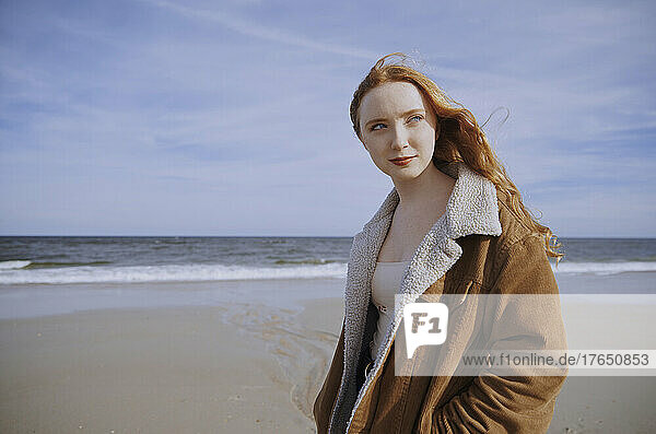 Redhead teenage girl wearing brown jacket standing at beach