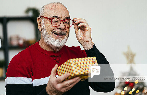 Smiling senior man wearing eyeglasses looking at gift box at home