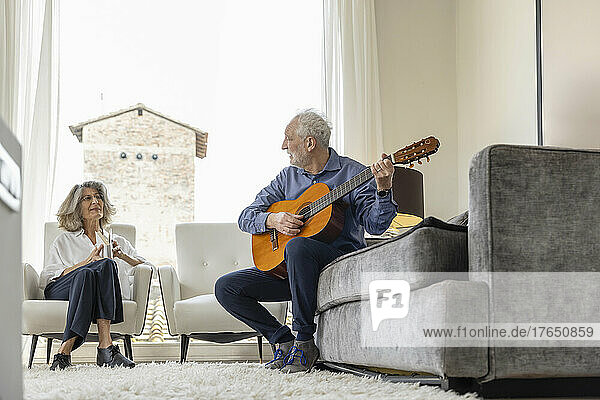 Senior woman looking at man playing classical guitar sitting on sofa at home