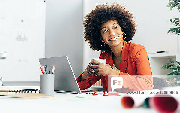 Smiling businesswoman holding coffee cup sitting with laptop at desk in office