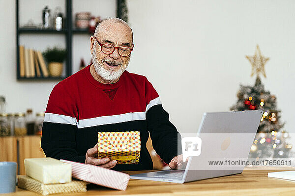 Senior man holding gift box using laptop at home