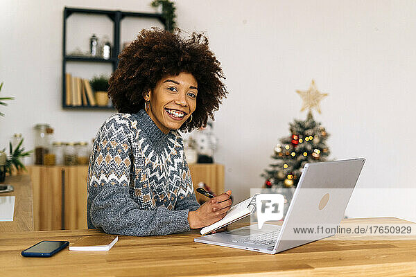Happy young woman with note pad and laptop sitting at table in living room