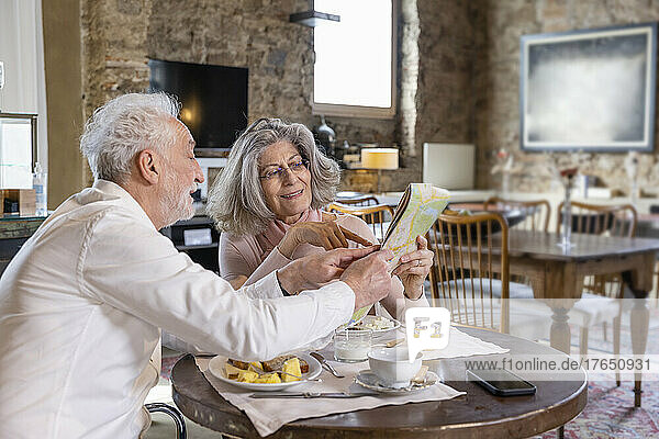 Senior man showing city map to woman sitting at breakfast table in boutique hotel