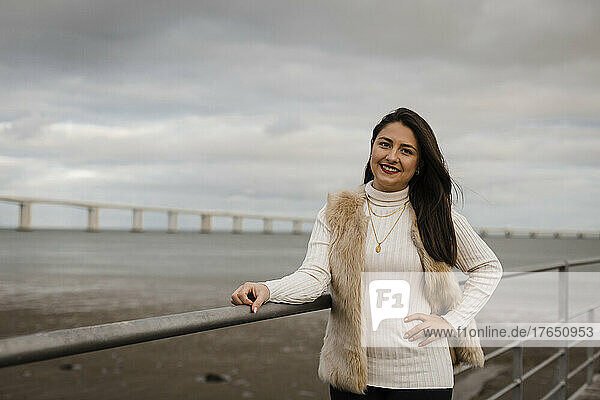 Smiling woman with hand on hip standing by railing in front of sea