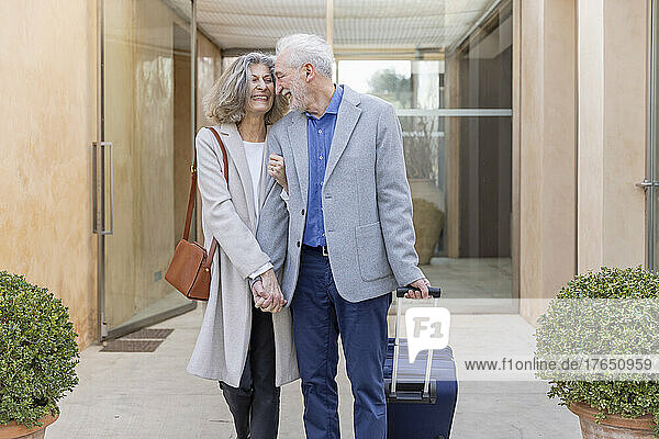 Cheerful senior couple with suitcase arriving at boutique hotel