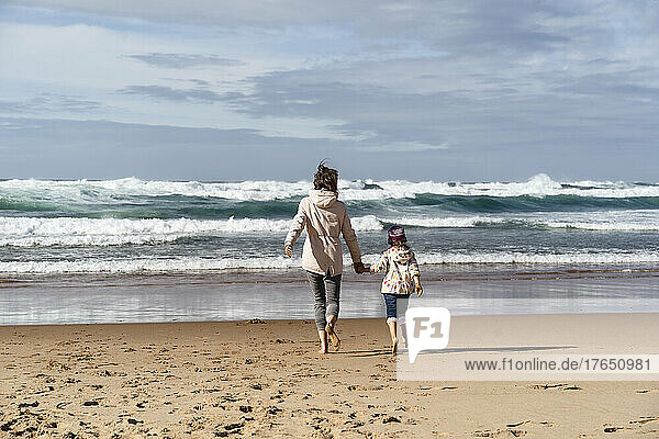 Mother and daughter walking towards waves splashing at beach on sunny day