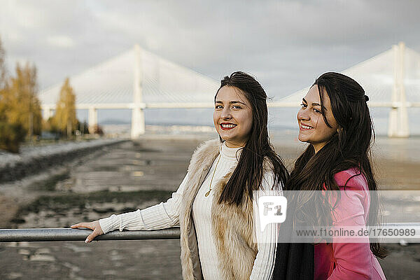Happy sisters standing by railing in front of bridge